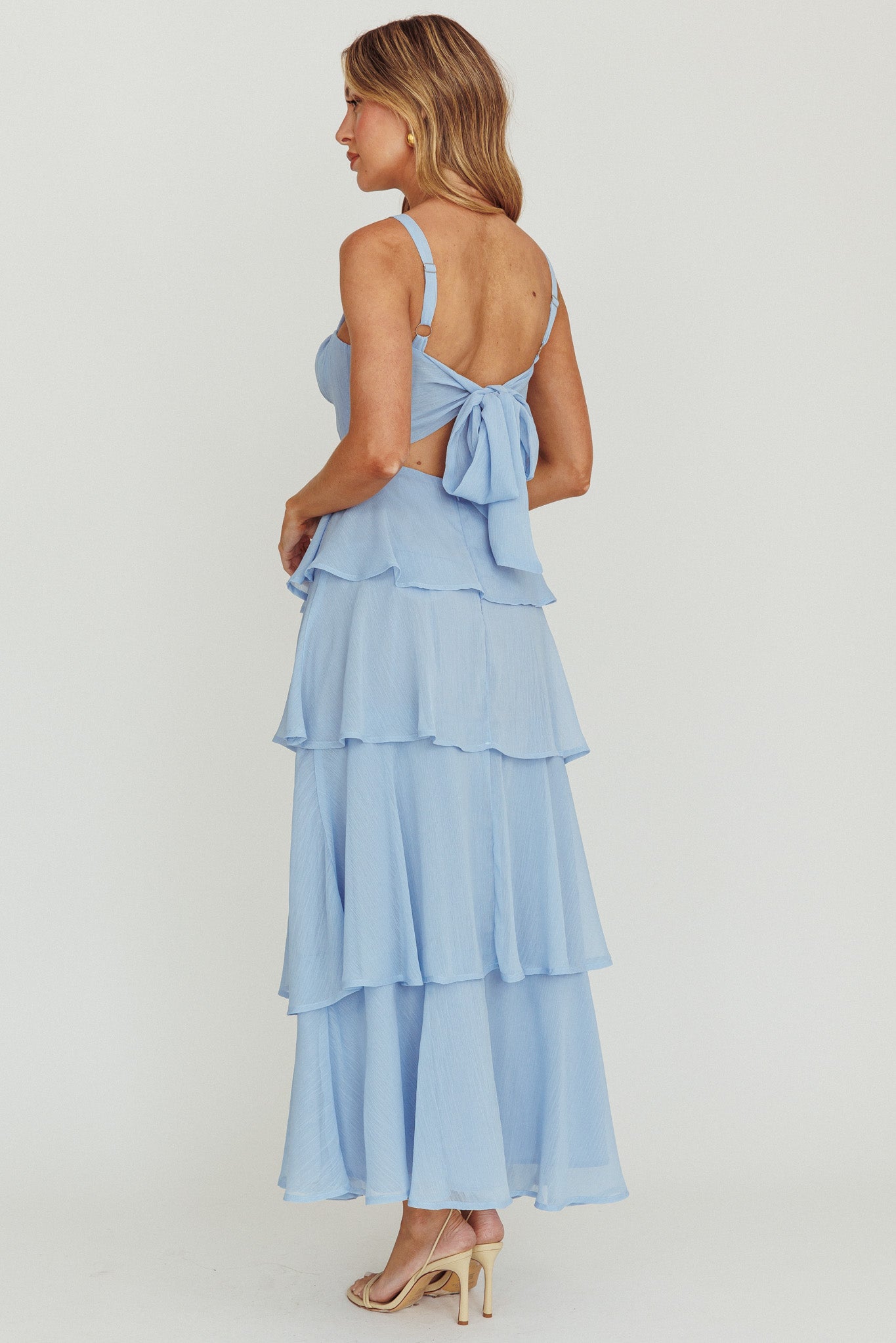 A woman is wearing a light blue, tiered maxi dress with a tie-back detail. She stands in a neutral studio setting, showcasing the dress's flowing layers and her strappy heels.