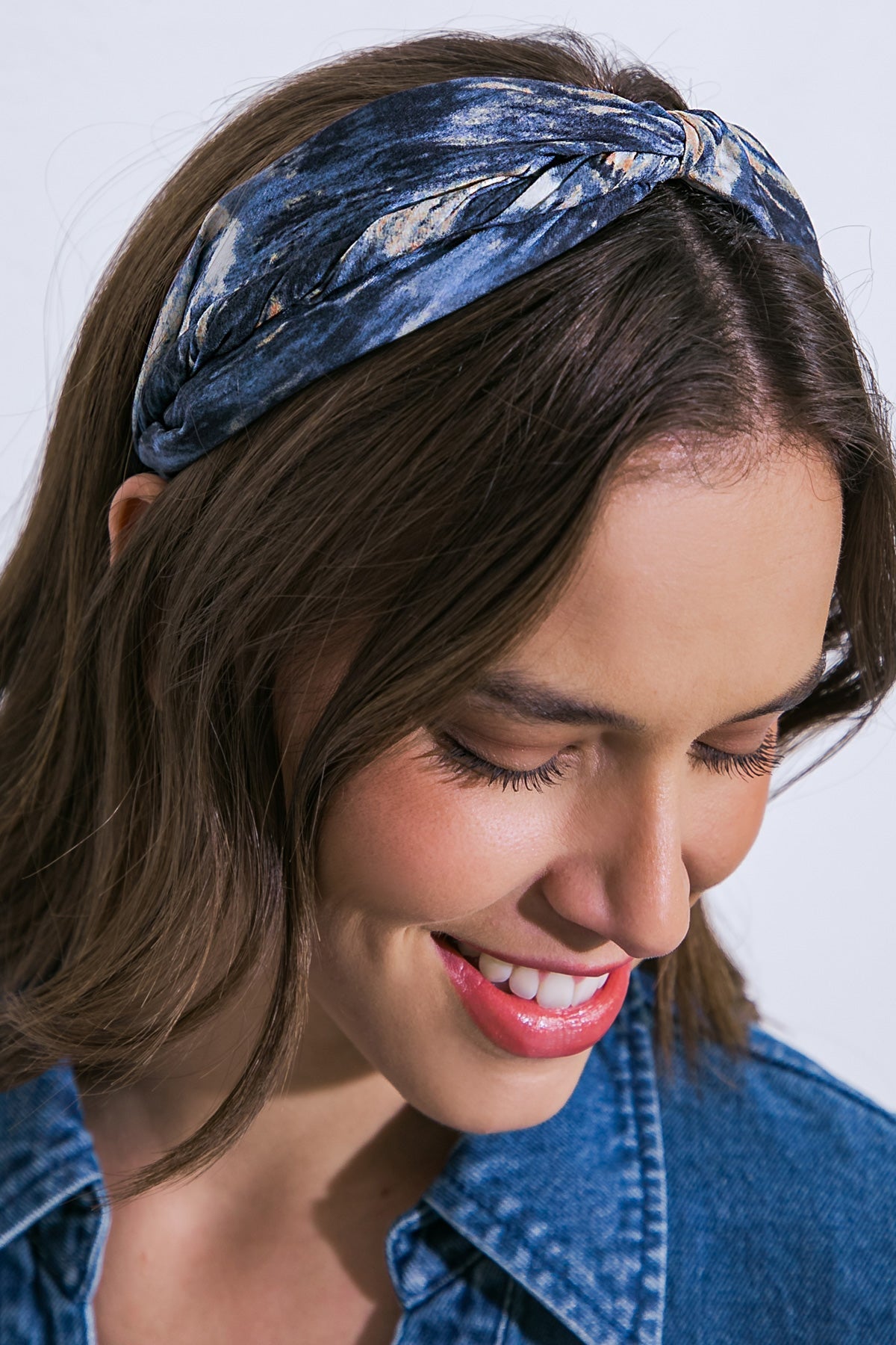 A woman with a blue patterned headband smiles softly, looking downward. She wears a denim jacket, and her hair cascades around her face against a light background.