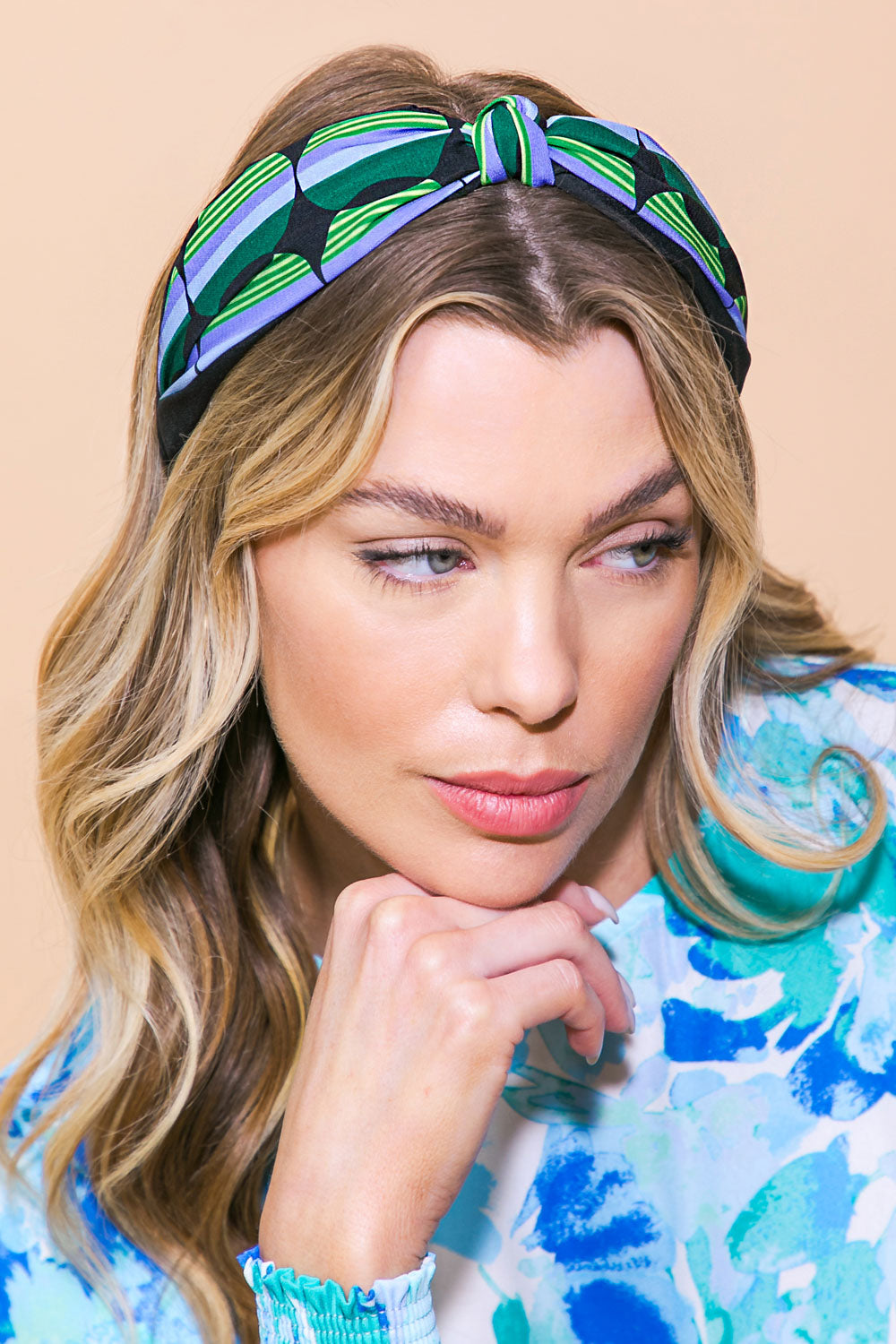 A woman with long, wavy hair is resting her chin on her hand, wearing a colorful patterned headband and a blue floral top, set against a neutral background.