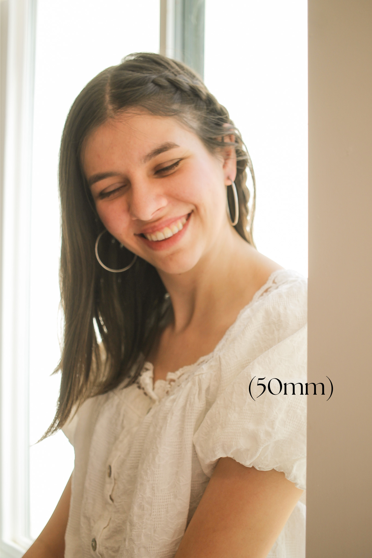 A young woman with long hair smiles gently, gazing downward while standing near a window. She wears a white, textured blouse and has hoop earrings. Soft natural light illuminates the scene.