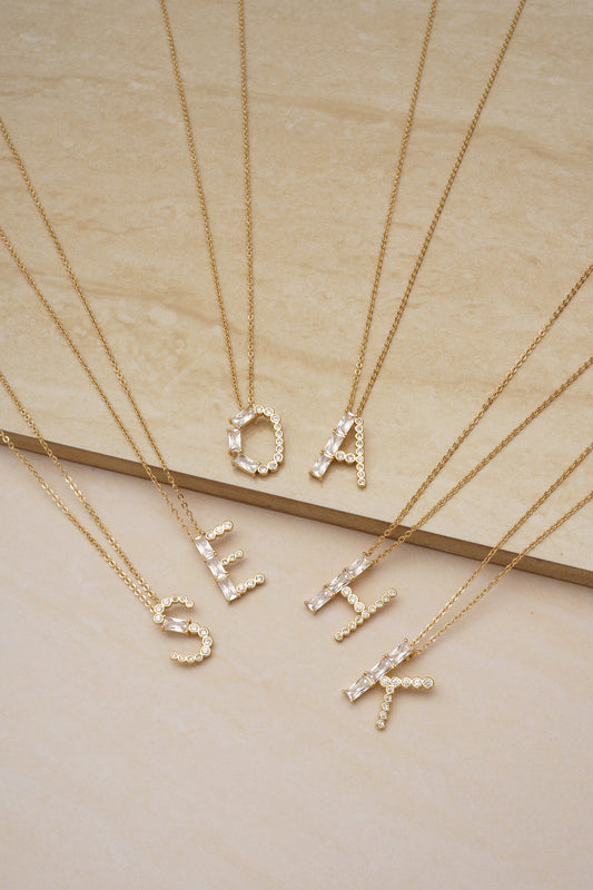 Gold necklaces featuring individual letters are arranged on a light-colored marble surface, each letter decorated with clear gemstones, showcasing a polished and elegant presentation.