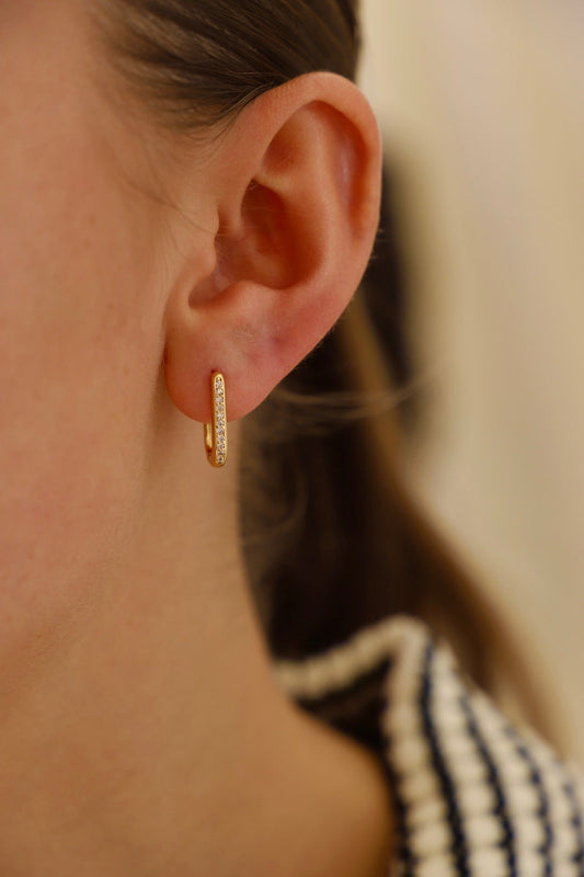 A gold hoop earring with a diamond-like embellishment hangs from a young woman's ear, partially obscured by her hair, set against a blurred, neutral-colored background.