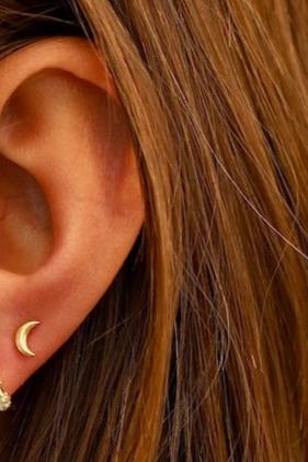 A close-up of an ear with a simple, gold crescent moon earring. The surrounding environment features long, brown hair partially obscuring the ear, creating a casual, stylish look.