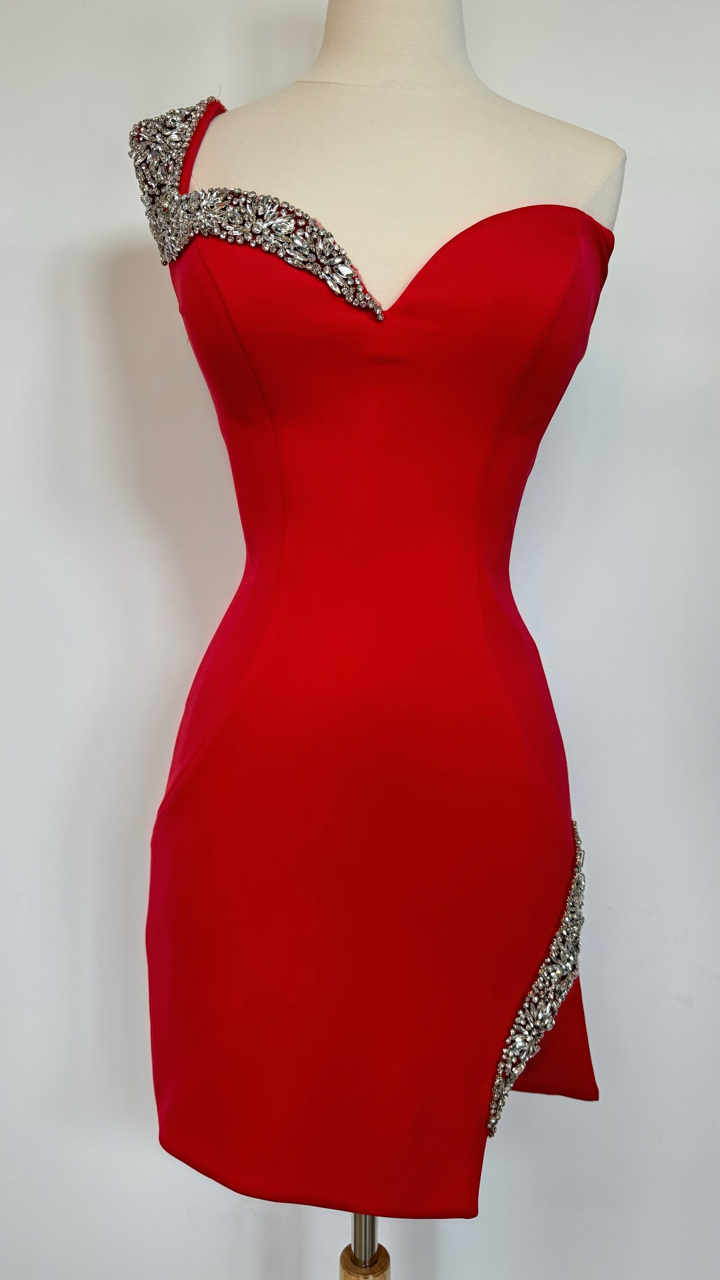 A red evening dress is displayed on a mannequin. It features a sweetheart neckline with a sparkling embellishment on one shoulder and along the hem, set against a plain background.