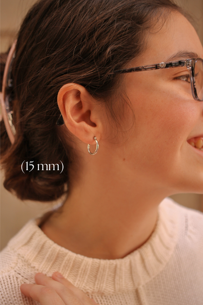 A woman with brown hair wears glasses and a cream-colored sweater, smiling. A small silver hoop earring is visible on her ear, labeled as (15 mm).