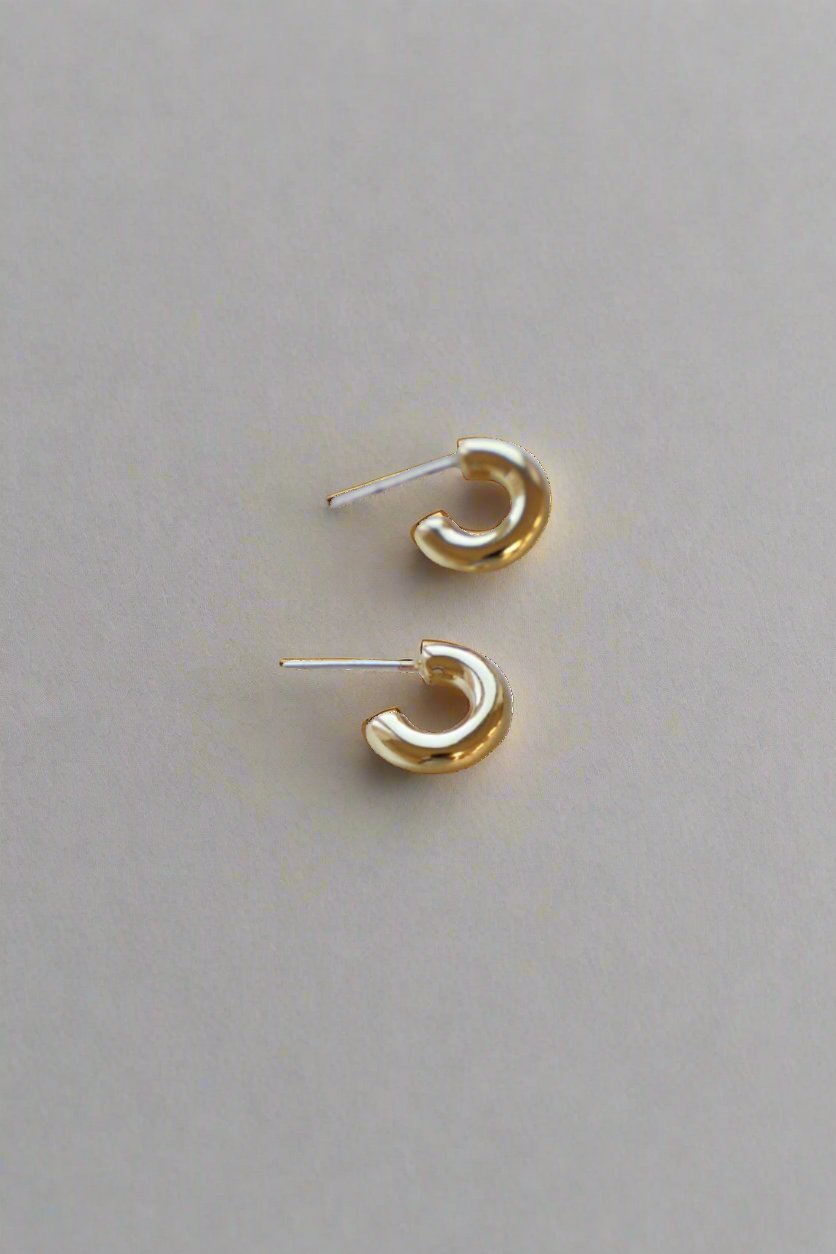 Two small, gold hoop earrings rest on a light-colored surface, showcasing their shiny, curved design. The earrings are oriented to display their circular shape and slender posts.