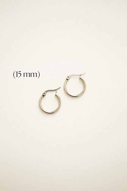 Two small silver hoop earrings, each 15 mm in diameter, rest on a light-colored, textured background. Their clasps are visible, indicating they are ready to be worn.