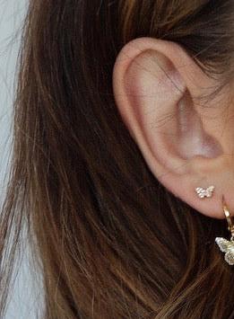 A close-up of an ear adorned with a small, delicate butterfly-shaped earring. The hair is styled loose, partially covering the ear, set against a neutral background.