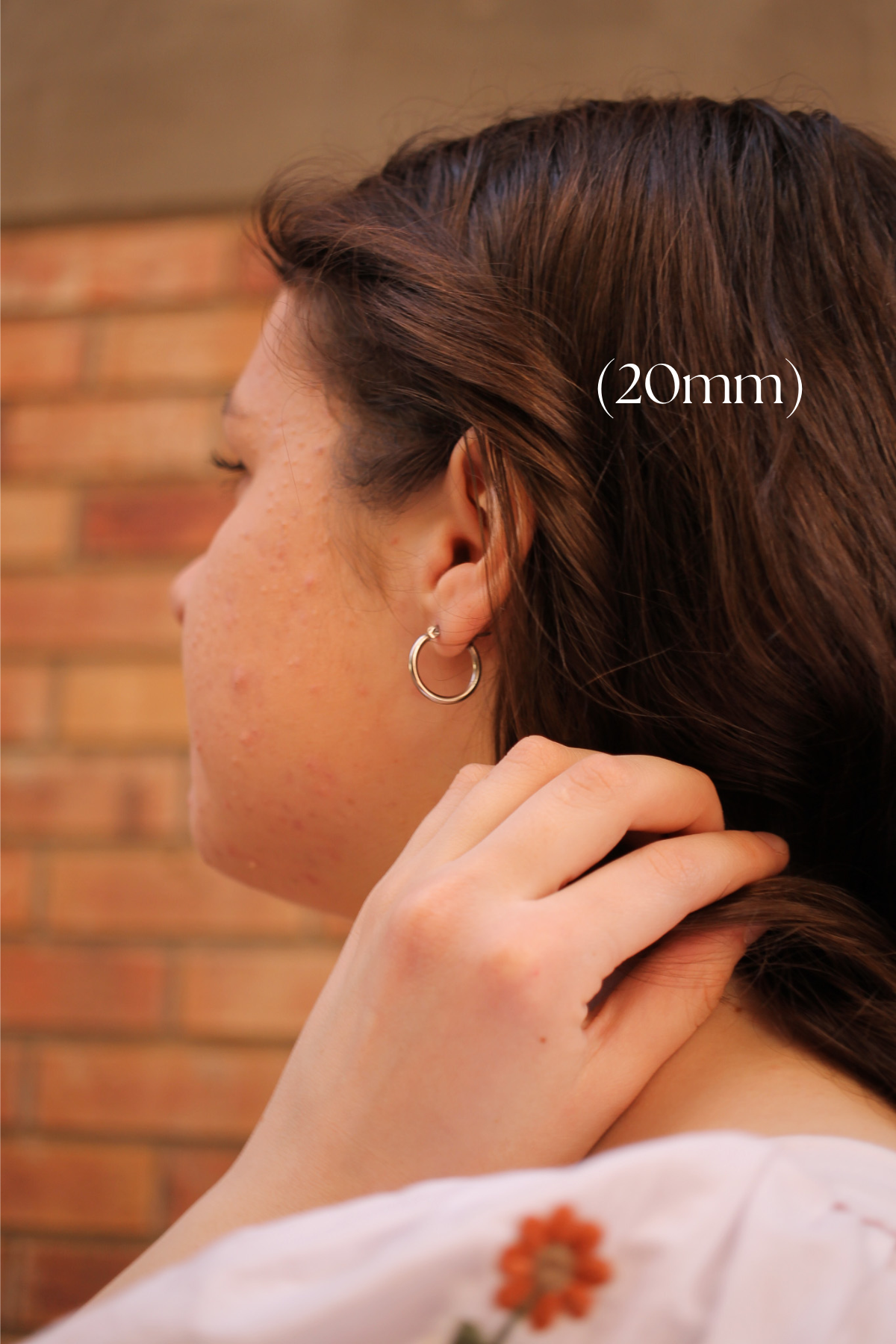 A young woman with shoulder-length brown hair gently tucks a strand behind her ear, showcasing a small hoop earring against a backdrop of textured brick walls. Text reads "(20mm)."