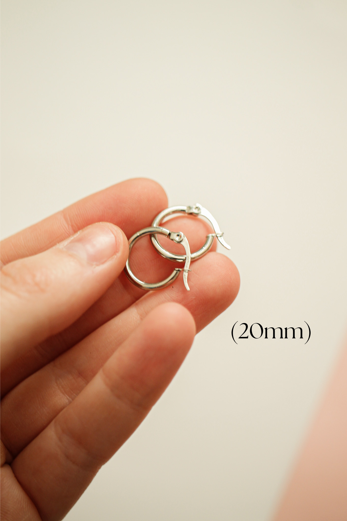 A hand holds two silver hoop earrings, displaying their circular shape and clasp mechanism. The background is softly blurred, emphasizing the earrings against a neutral tone. (20mm)