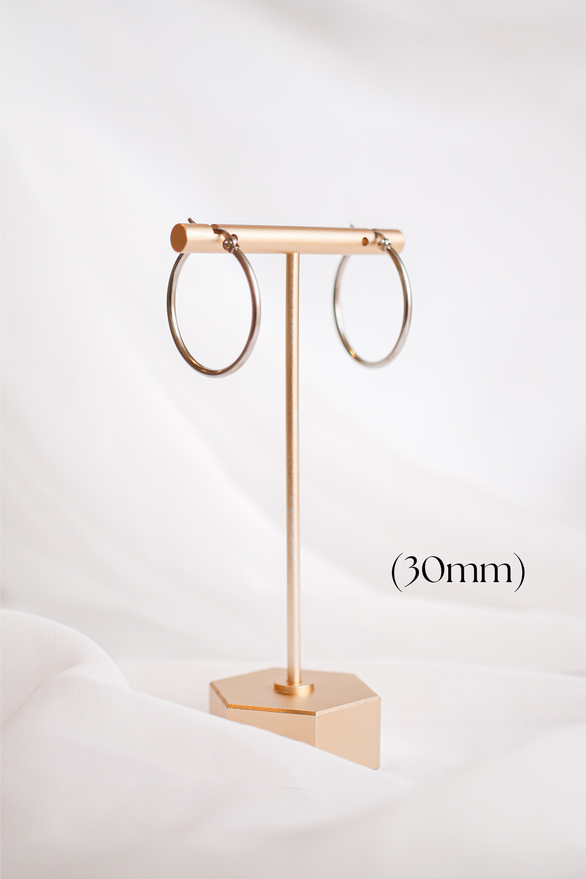 A pair of circular silver hoop earrings hangs on a gold display stand, set against a softly draped white background. (30mm) is noted below.