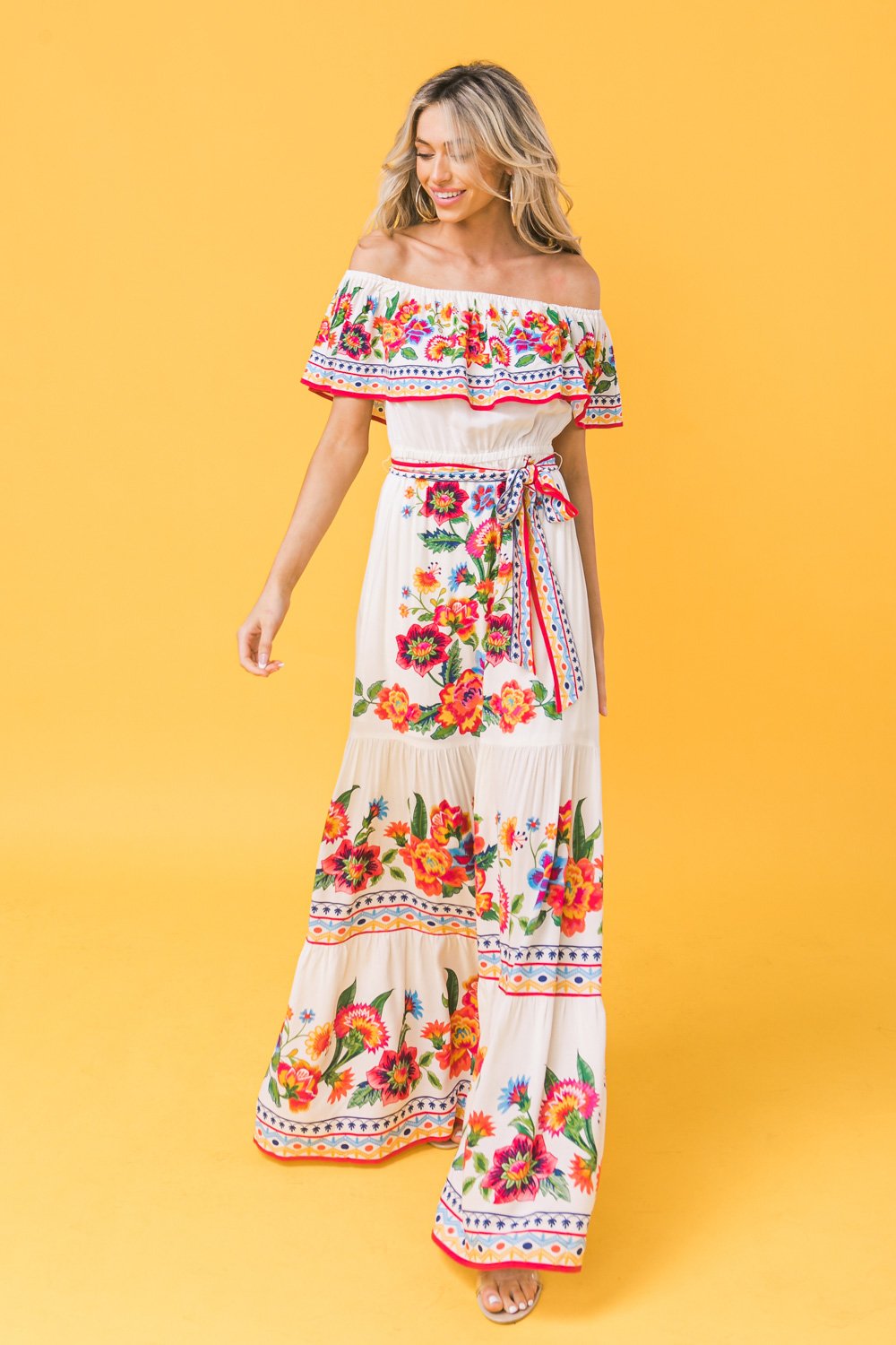 A woman wearing a colorful floral maxi dress with ruffled sleeves is smiling and walking. The background is a bright yellow, enhancing the vibrant colors of her outfit.