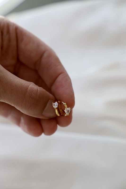 A hand holds a gold ring featuring two heart-shaped gemstones. It is positioned against a softly textured, light-colored background, suggesting a delicate and intimate setting.