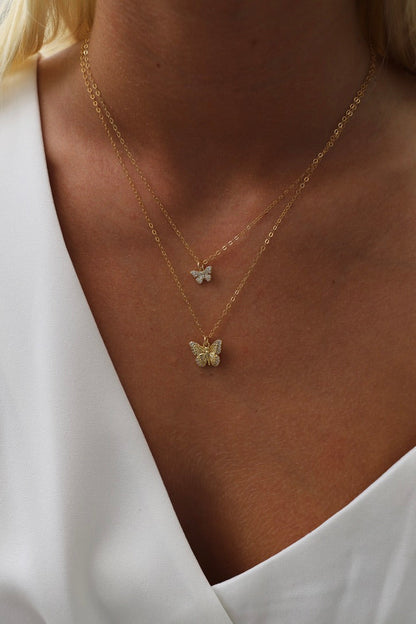 A close-up of a person's neck features two delicate gold necklaces. One has a small silver butterfly, and the other displays a larger, ornate gold butterfly, against a light fabric backdrop.