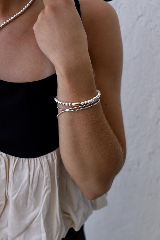 A hand adorned with multiple silver and gold bracelets is resting against a bare arm, with the person wearing a black top and a light-colored, ruffled garment, set against a textured white background.