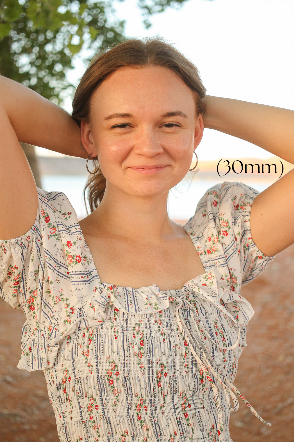 A young woman is smiling while pulling her hair back, wearing a floral-patterned dress. The background features a blurred outdoor setting with natural light and water visible. Text in the image reads "(30mm)".