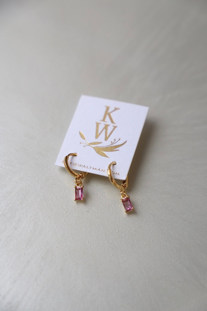 Gold hoop earrings with pink rectangular gemstones hang from a white card featuring â€œKWâ€ and a simple illustration. The setting is a reflective, light-colored surface.