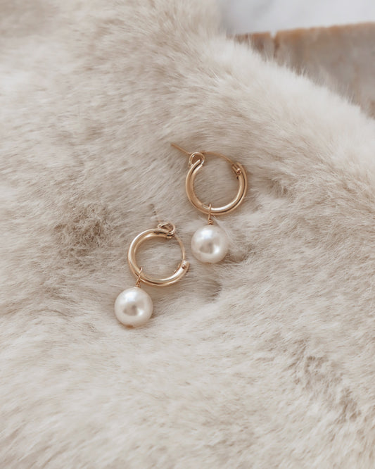 Gold hoop earrings with attached white pearls rest on a soft, textured cream-colored surface, creating an elegant display that highlights the jewelry's design and shine.