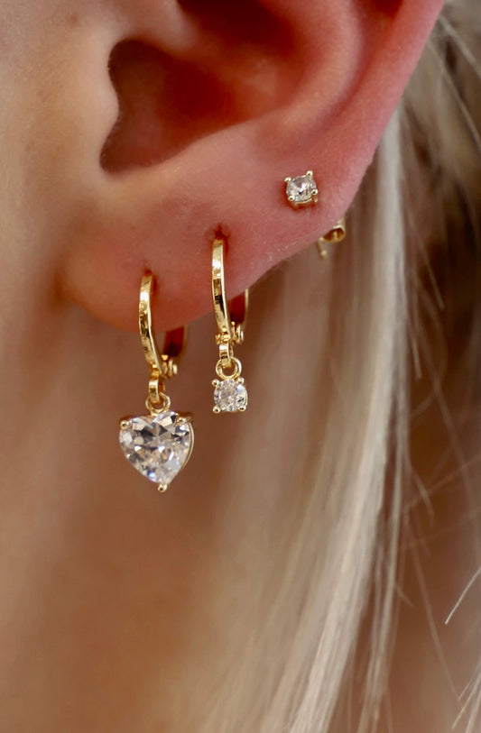 Golden earrings dangle from a lobe, one shaped like a heart with a large clear stone and another smaller stone. The background features lightly colored hair, adding a soft touch.