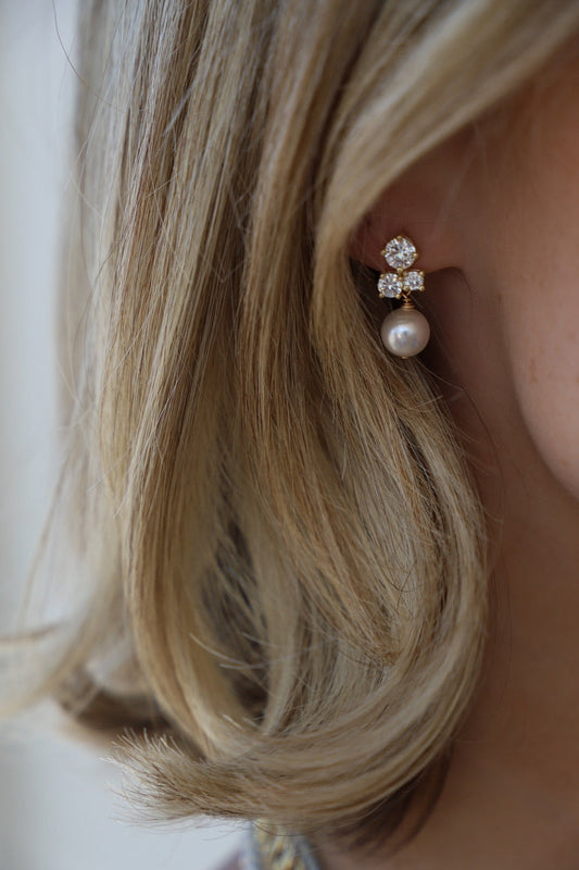 A pair of elegant earrings, featuring a pearl and sparkling stones, adorns a woman's ear, framed by cascading blonde hair, set against a neutral background.