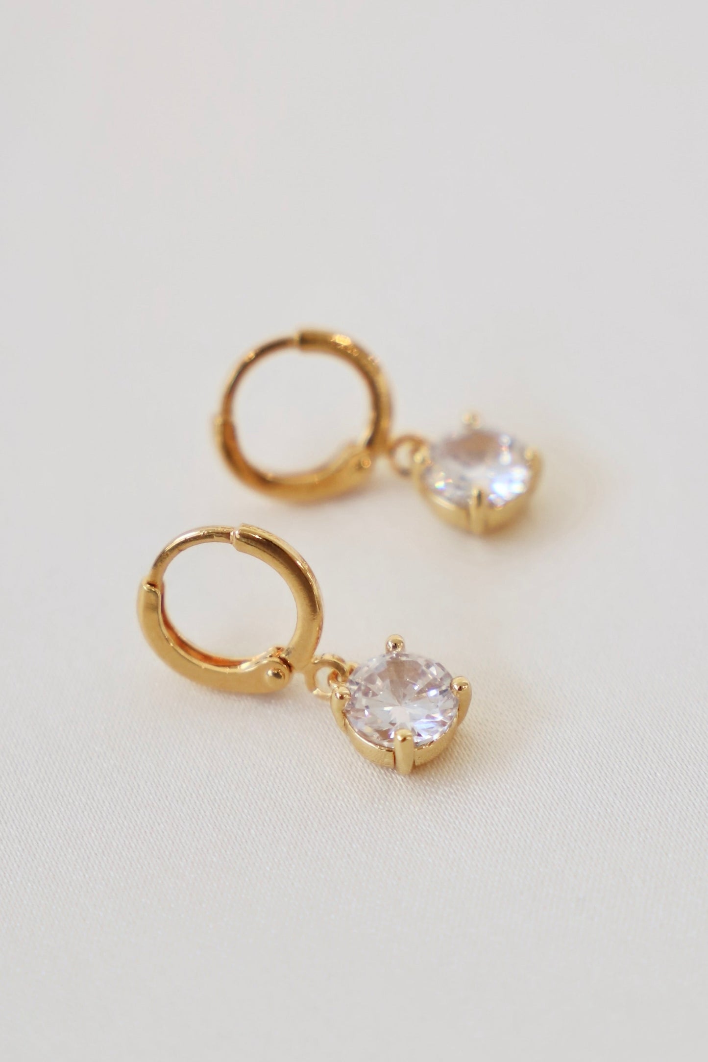 Gold hoop earrings with a dangling crystal feature shine against a soft, light-colored fabric surface, adding elegance and sparkle to the jewelry display.