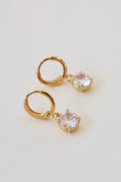 Gold hoop earrings with a dangling crystal feature shine against a soft, light-colored fabric surface, adding elegance and sparkle to the jewelry display.