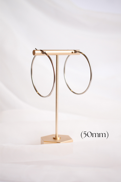 Two metallic hoop earrings hang on a simple gold display stand against a soft, white fabric background. The earrings are labeled with the diameter size, 50mm.