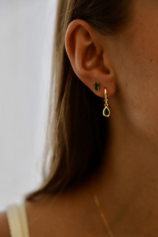 A close-up of a woman's ear showcases a gold hoop earring with a green gemstone, alongside a smaller green stud earring, set against a softly blurred background.