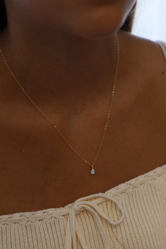 A delicate gold necklace with a small opal pendant rests on the neckline of a person wearing a light, textured garment with a tie detail, set against a softly lit background.