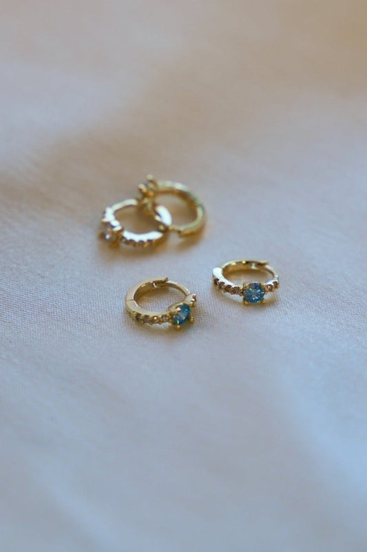 Four pairs of gold hoop earrings rest on a soft, light-colored fabric. Two pairs feature small blue stones, while the others are plain gold, showcasing intricate designs.