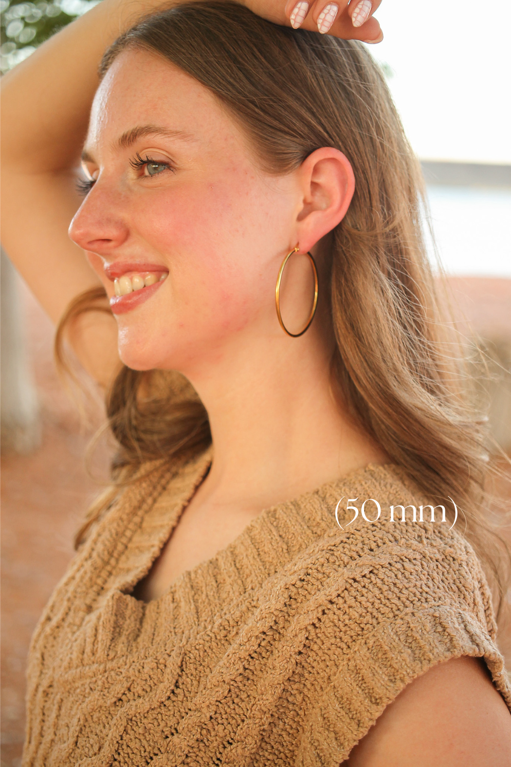 A young woman smiles, wearing large gold hoop earrings and a textured brown sweater, with her hand resting above her head against a soft, blurred outdoor background. Text reads "(50 mm)".