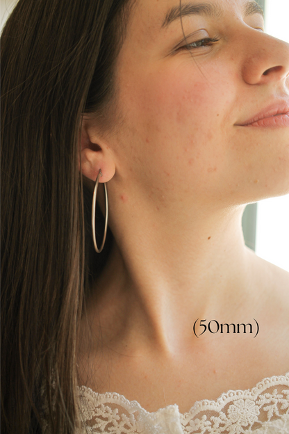 A young woman wearing a large hoop earring tilts her head slightly, smiling softly. She has long hair and is dressed in a lace-top garment with natural lighting from a window. (50mm)