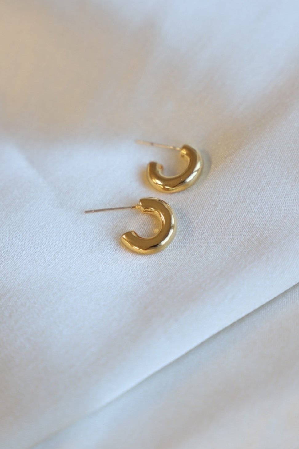 Two gold hoop earrings rest on a light-colored fabric surface. The earrings feature a curved, polished design, showcasing a shiny finish against the soft backdrop.