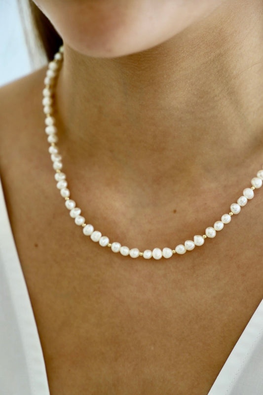 A pearl necklace rests on a person’s neck, worn over a white garment, creating a soft and elegant appearance in a well-lit setting.