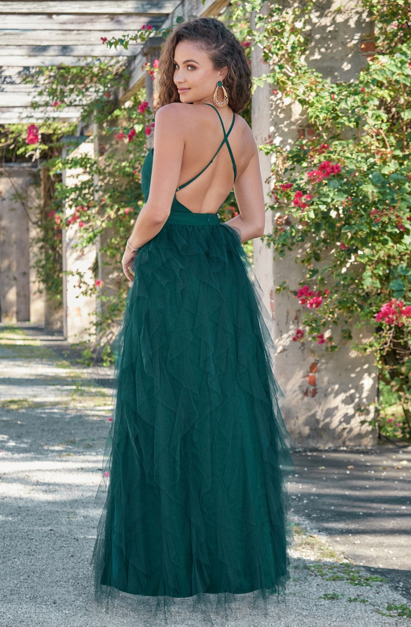 A woman in a flowing, green gown with ruffled tulle stands with her back towards the camera, surrounded by lush greenery and vibrant flowers in a picturesque outdoor setting.