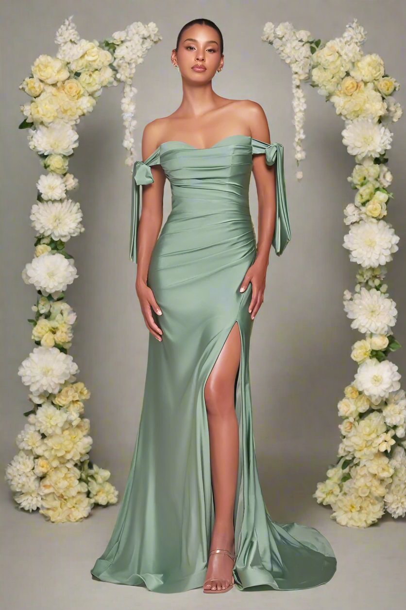 A model poses gracefully in a silky, off-shoulder, light green gown with a thigh-high slit, surrounded by floral arrangements of white and yellow blooms against a soft gray backdrop.