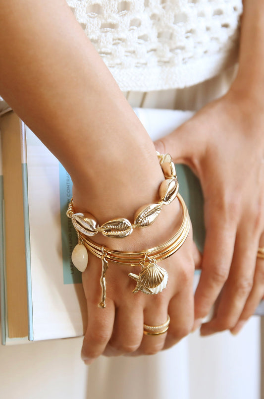 A hand adorned with multiple gold bracelets, showcasing a shell motif and charms, is holding a book. The background features a light, textured fabric, enhancing the delicate aesthetics.