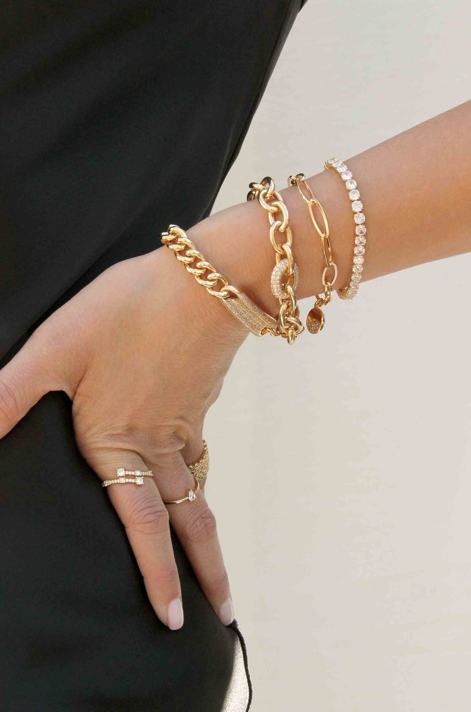 A hand adorned with multiple gold and diamond bracelets and rings holds a black garment, set against a light background that creates a soft, elegant ambiance.