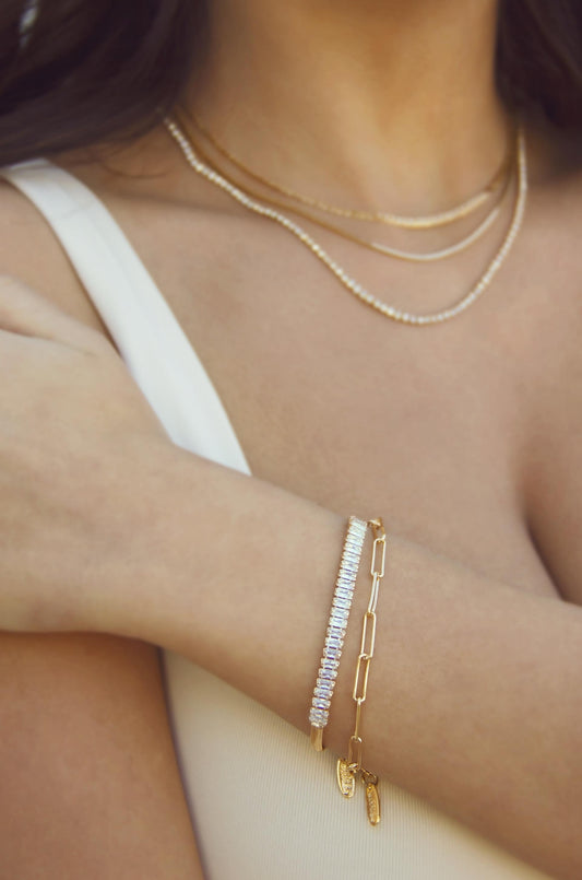 A delicate wrist adorned with two stylish bracelets, one featuring sparkling stones and the other a gold chain with charms, set against bare skin and minimalistic attire.