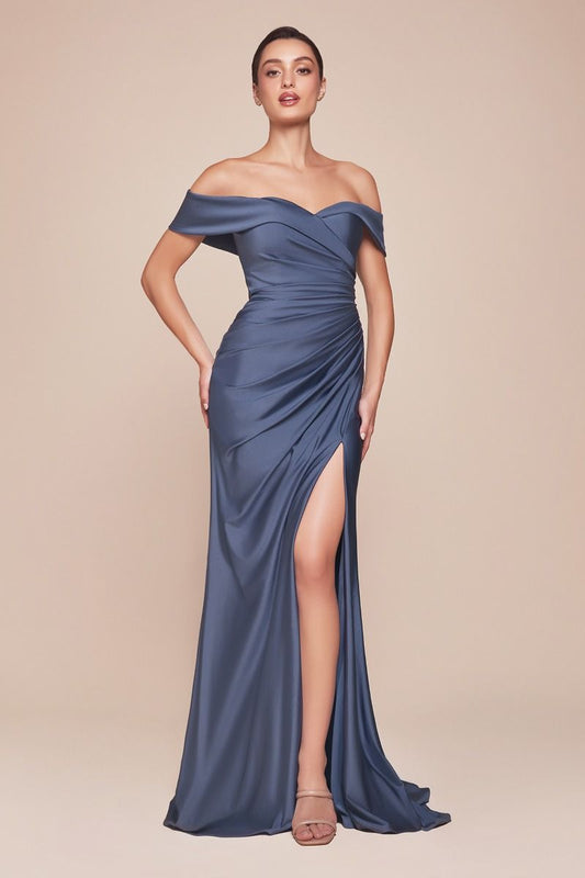 A woman stands confidently in a floor-length, off-shoulder blue gown with a thigh-high slit, showcasing elegance. The background is a soft neutral tone, enhancing the dress's features.