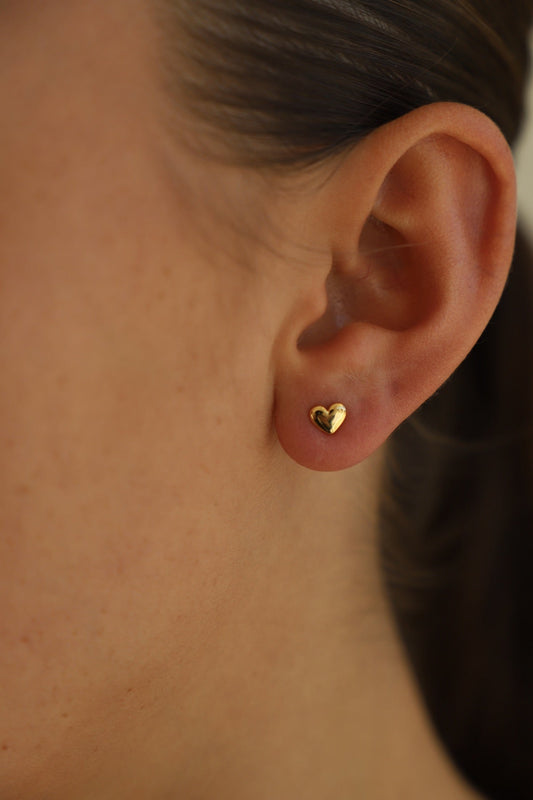 A small, heart-shaped gold earring is worn on a woman's ear. The skin is smooth and lightly freckled, with the hair tucked back, highlighting the earring's shine against a neutral background.