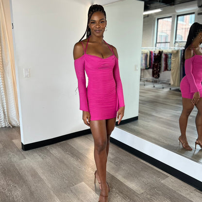 A woman stands confidently in a form-fitting, pink dress with long sleeves and off-shoulder cutouts. She poses in a clothing boutique, with dresses visible on racks behind her.