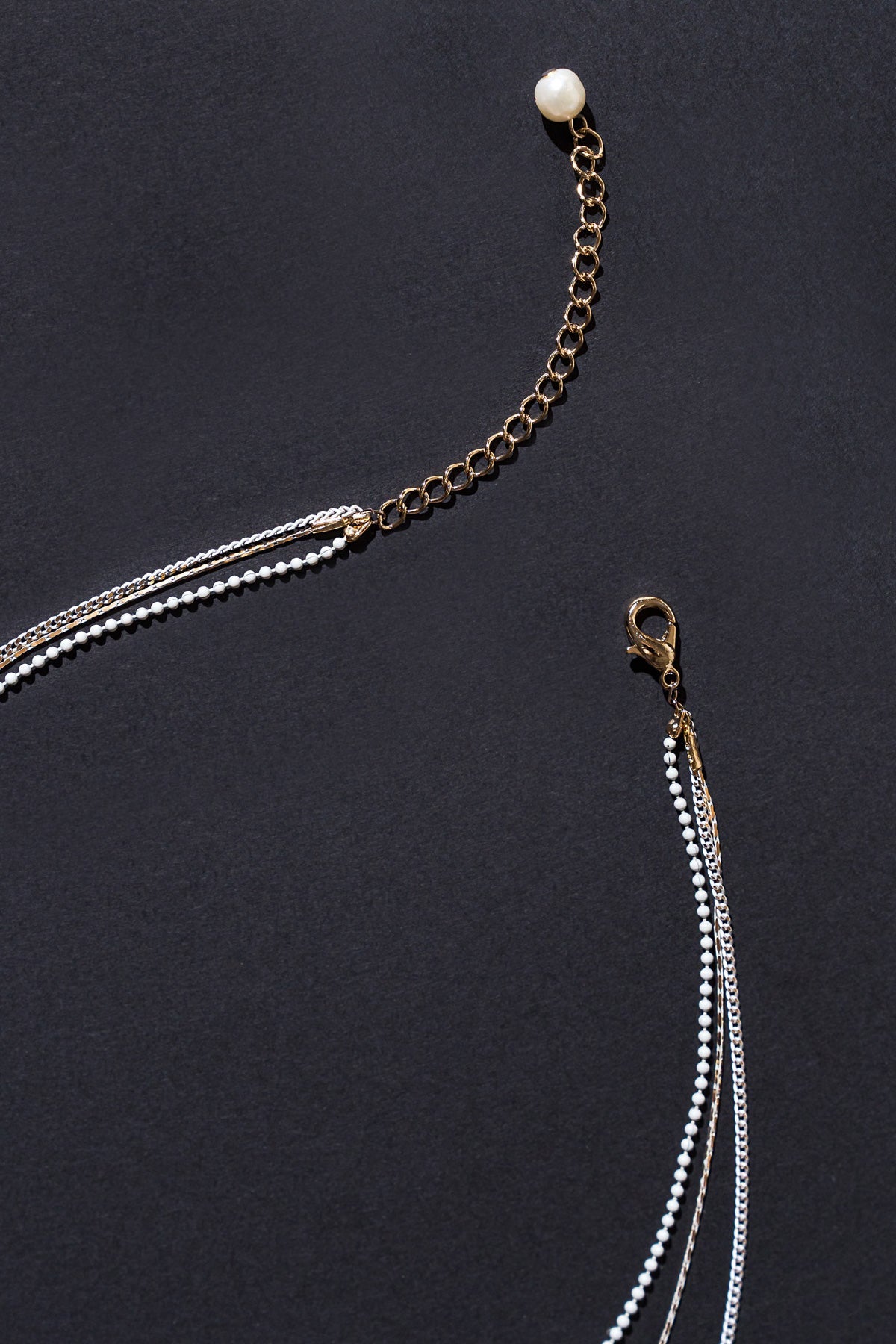 A gold chain necklace with a white bead and several strands of varying lengths, featuring small white beads and metallic chains, is laid out against a dark background.