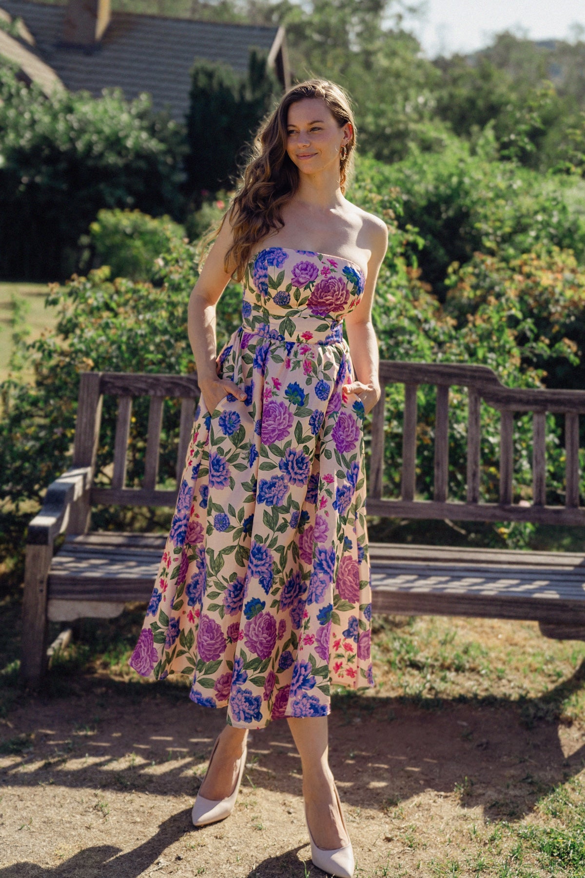 A woman in a floral-patterned strapless dress stands confidently, hands in pockets, on a grassy area near a wooden bench, surrounded by lush greenery and a sunny atmosphere.