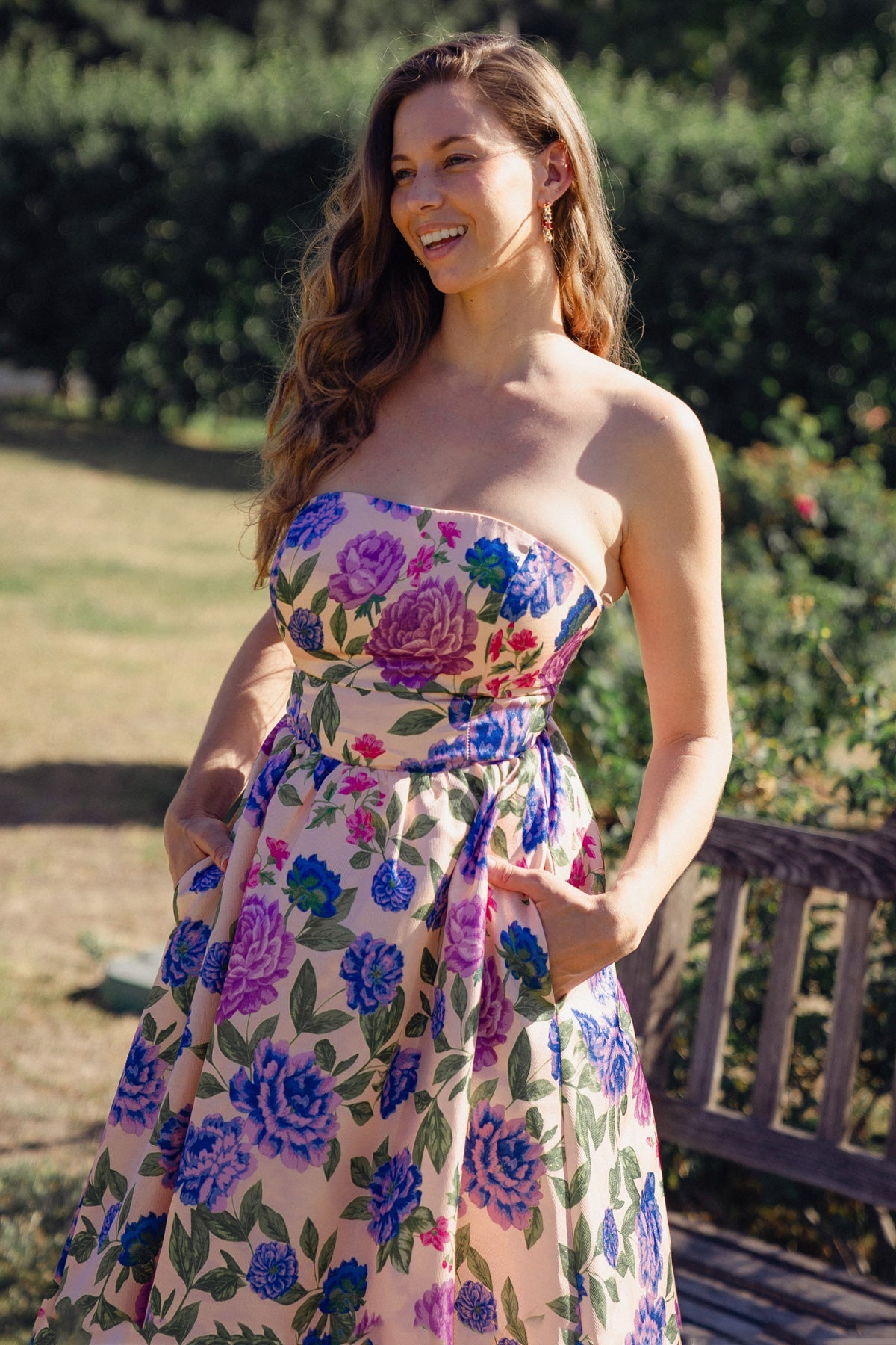 A woman stands smiling while wearing a strapless dress adorned with colorful floral patterns. She has her hands in the pockets, surrounded by greenery and a rustic wooden bench.