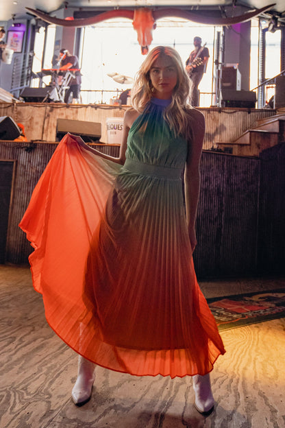 A woman in a gradient pleated dress stands confidently, holding her flowing skirt. Behind her, musicians are performing in a lively venue, illuminated by ambient lighting.