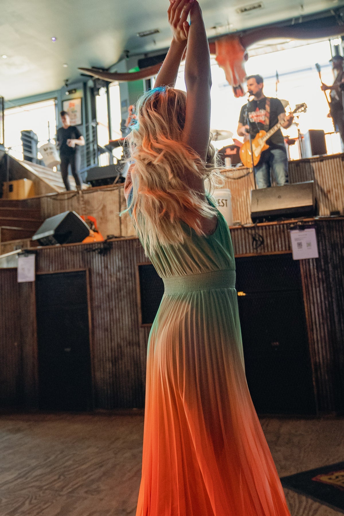 A woman wearing a colorful, flowing dress dances with her arms raised in celebration. In the background, a band performs in a lively venue, surrounded by music equipment and bar decor.