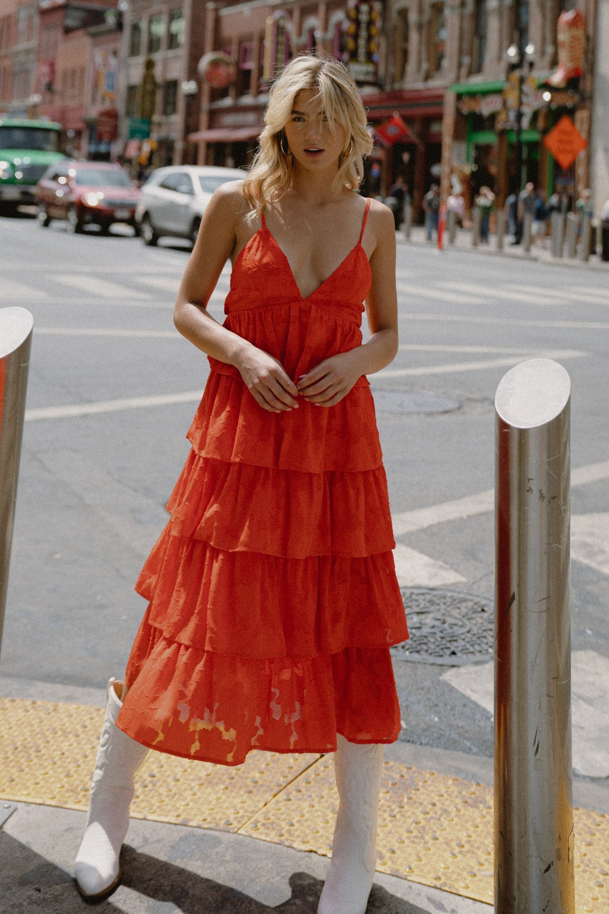 A woman wearing a layered red dress and white boots stands on a city street, looking directly at the camera, with various cars and colorful buildings in the background.