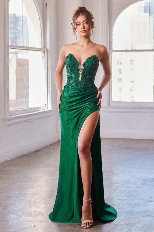 A woman stands confidently in a vibrant green evening gown with intricate lace detailing, displaying a high slit that reveals her leg, against a softly lit, urban backdrop with large windows.