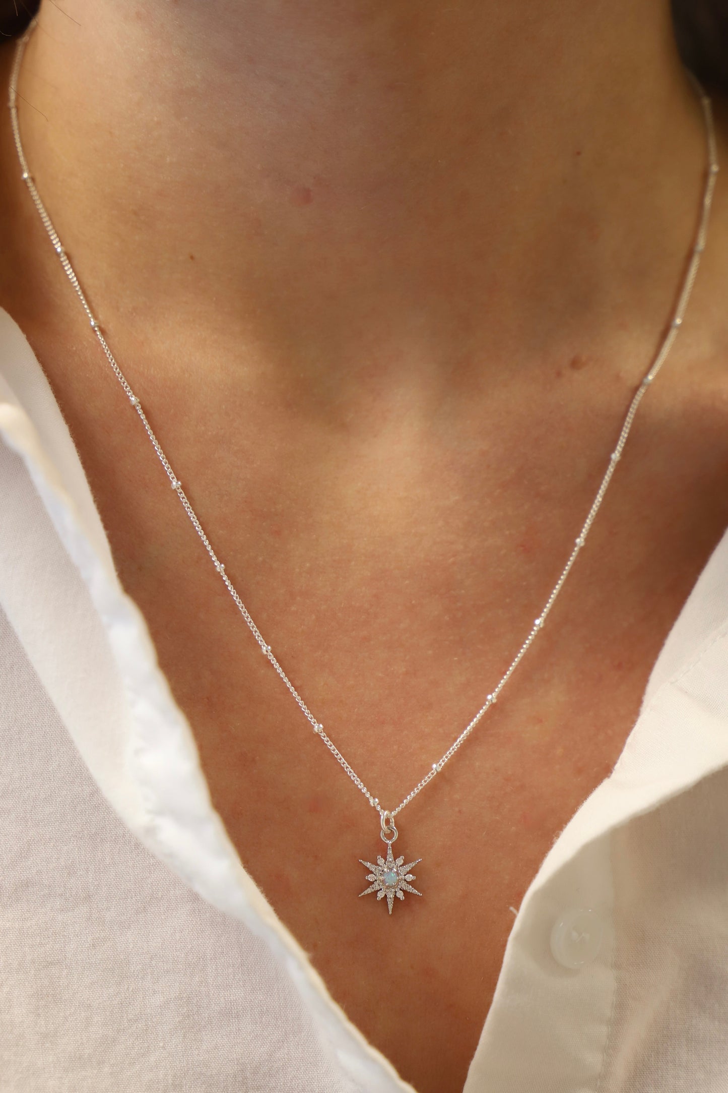 A silver chain necklace with a star-shaped pendant adorned with small gemstones rests on a person's neck, contrasting against their skin and a white shirt.